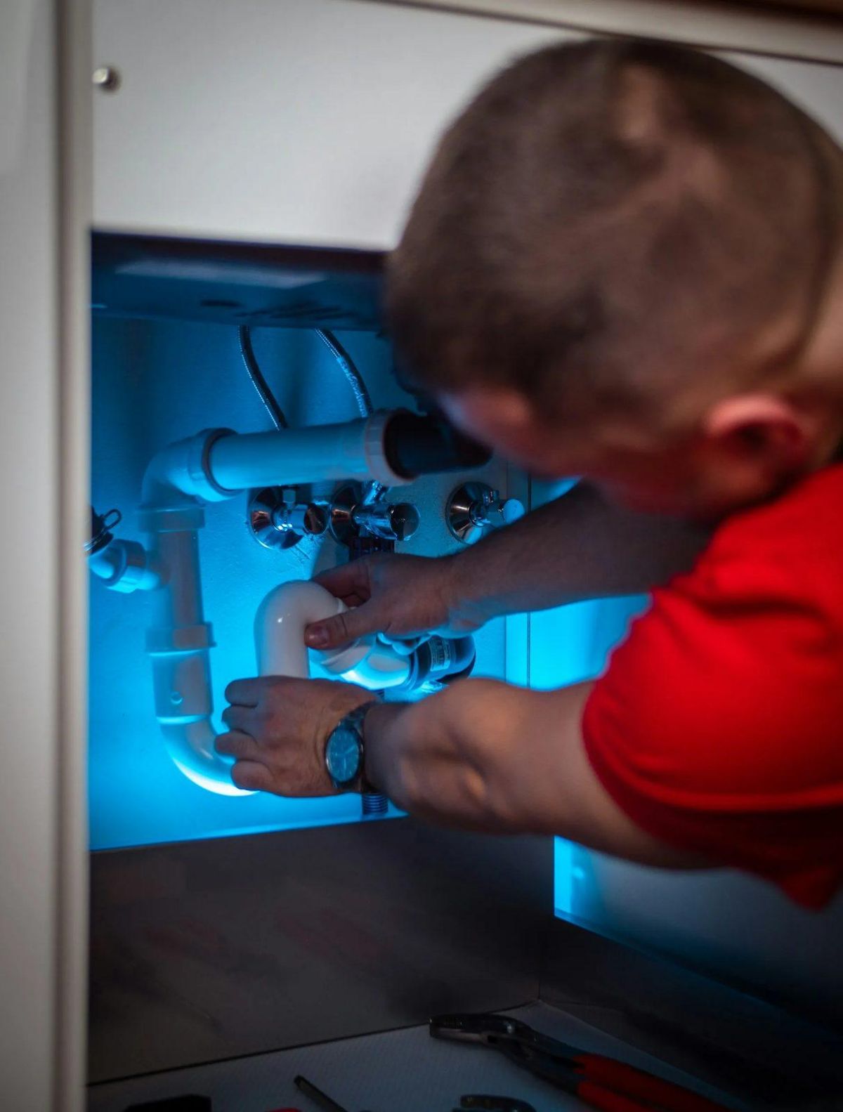 Plumber in a red shirt fitting white drain pipes under a sink