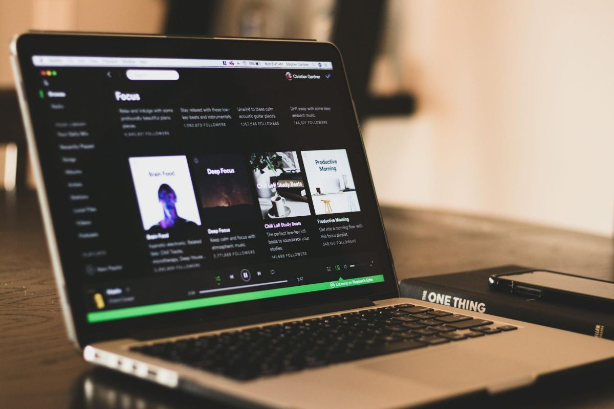 Laptop on a dark desk showing a music app beside a notebook