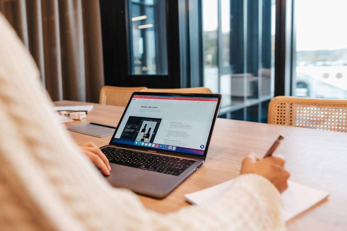 Person holding a pen while working on a laptop at a wooden table