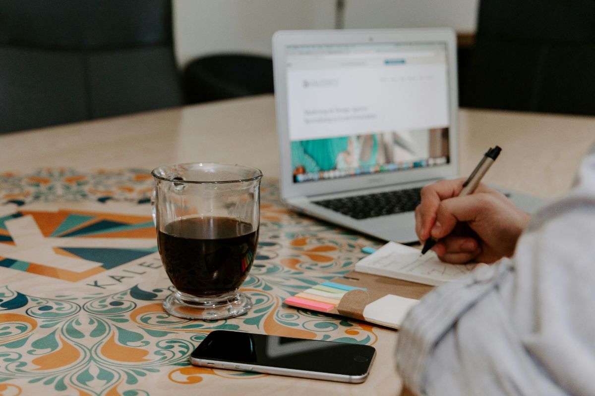 Person writing in a notepad next to a laptop, a phone and a glass of coffee