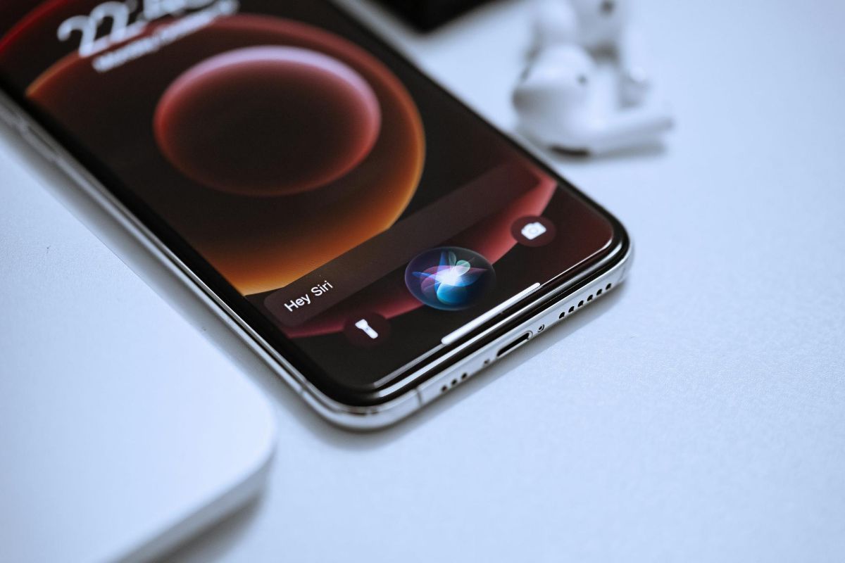 Phone on a white desk showing the Hey Siri screen next to an earbuds case